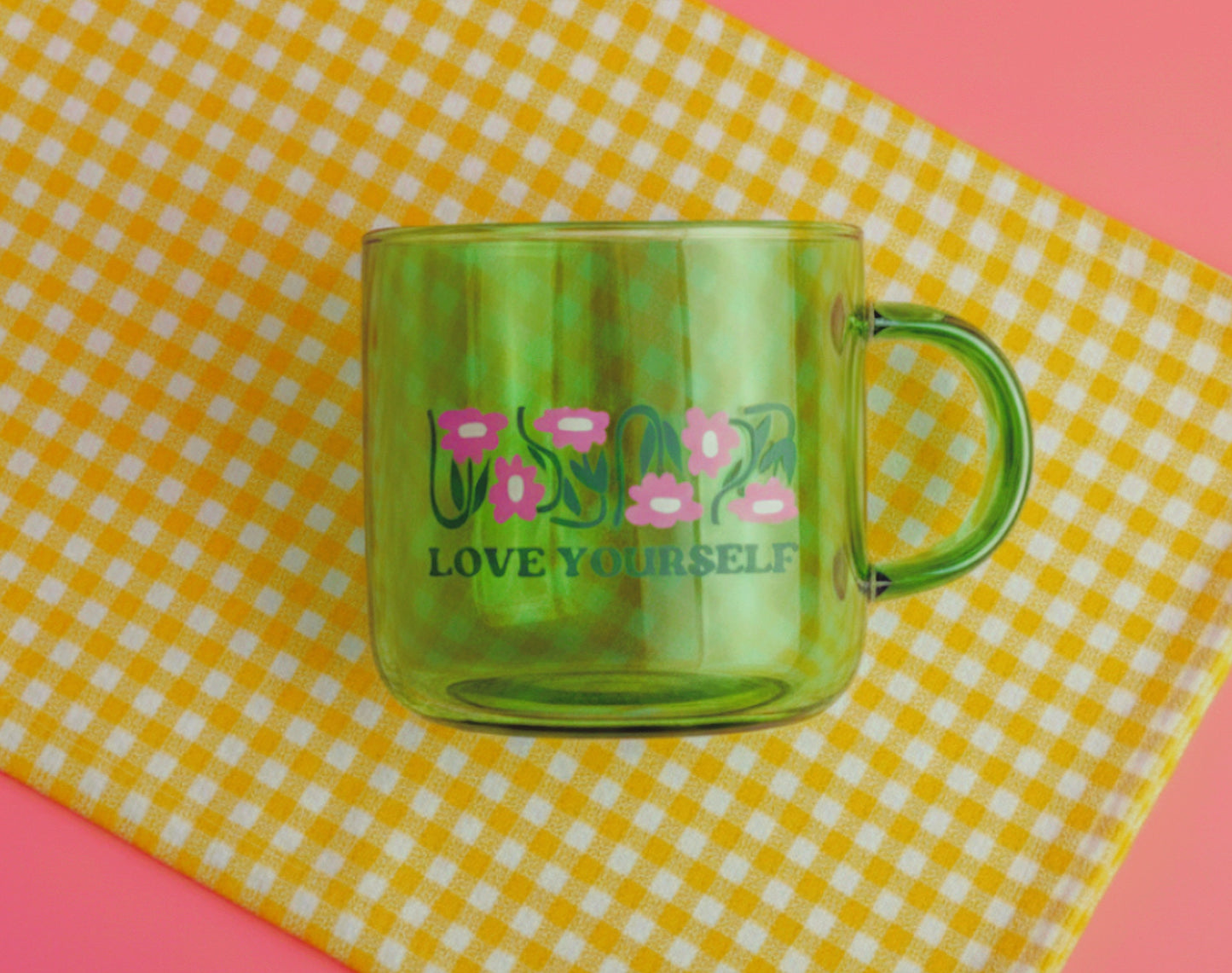 Green mug with 'LOVE YOURSELF' text and floral design on a yellow and white checkered tablecloth.