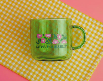 Green mug with 'LOVE YOURSELF' text and floral design on a yellow and white checkered tablecloth.